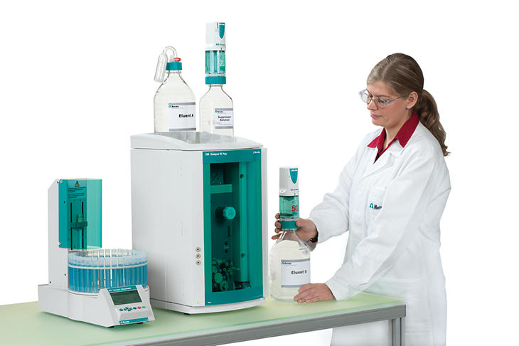 Female scientist in a lab coat and safety glasses operating Metrohm ion chromatography equipment, including a 930 Compact IC Flex instrument with eluent bottles labeled 'Eluent B' and 'Suppressor' on top. The scientist is preparing a sample next to the system in a well-lit laboratory