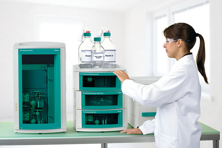 Female scientist in a lab coat and safety glasses operating Metrohm ion chromatography equipment, including an 940 Professional IC Vario instrument with eluent bottles labeled 'Eluent A' and 'Suppressor' on top, in a modern, well-lit laboratory