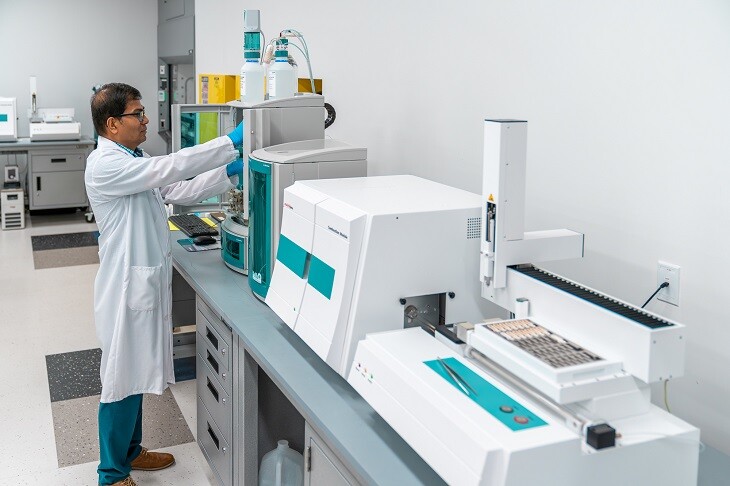 Male scientist in a lab coat working in a modern laboratory with various analytical instruments, including Metrohm ion chromatography equipment. The scientist is adjusting settings on the IC system, which is lined up with other advanced analytical devices on a long lab bench in a clean, well-organized environment